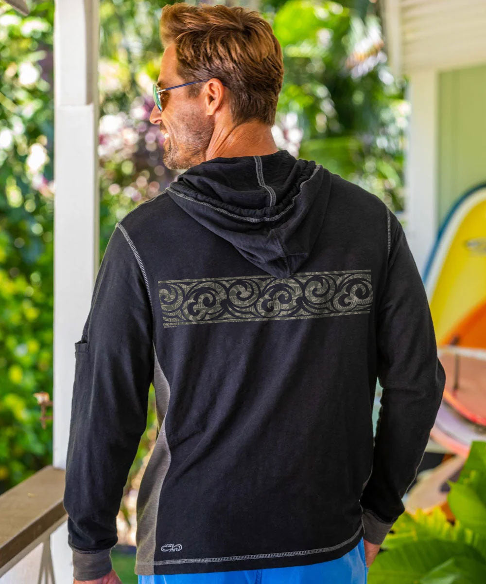 A man viewed from behind wearing a black long-sleeve hooded shirt with a decorative gray scroll pattern across the upper back, standing on a porch with green foliage in the background. View 3