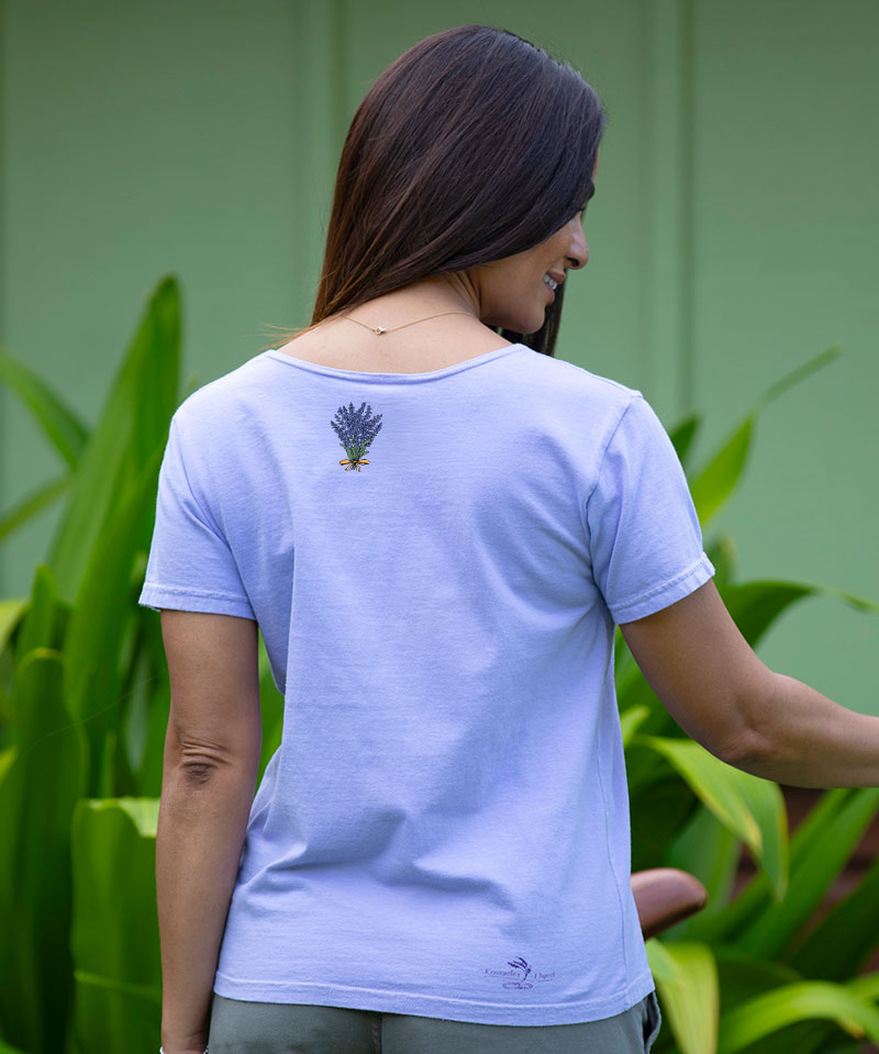 Woman wearing a light purple t-shirt with a logo on the back, standing in front of green foliage. View 3