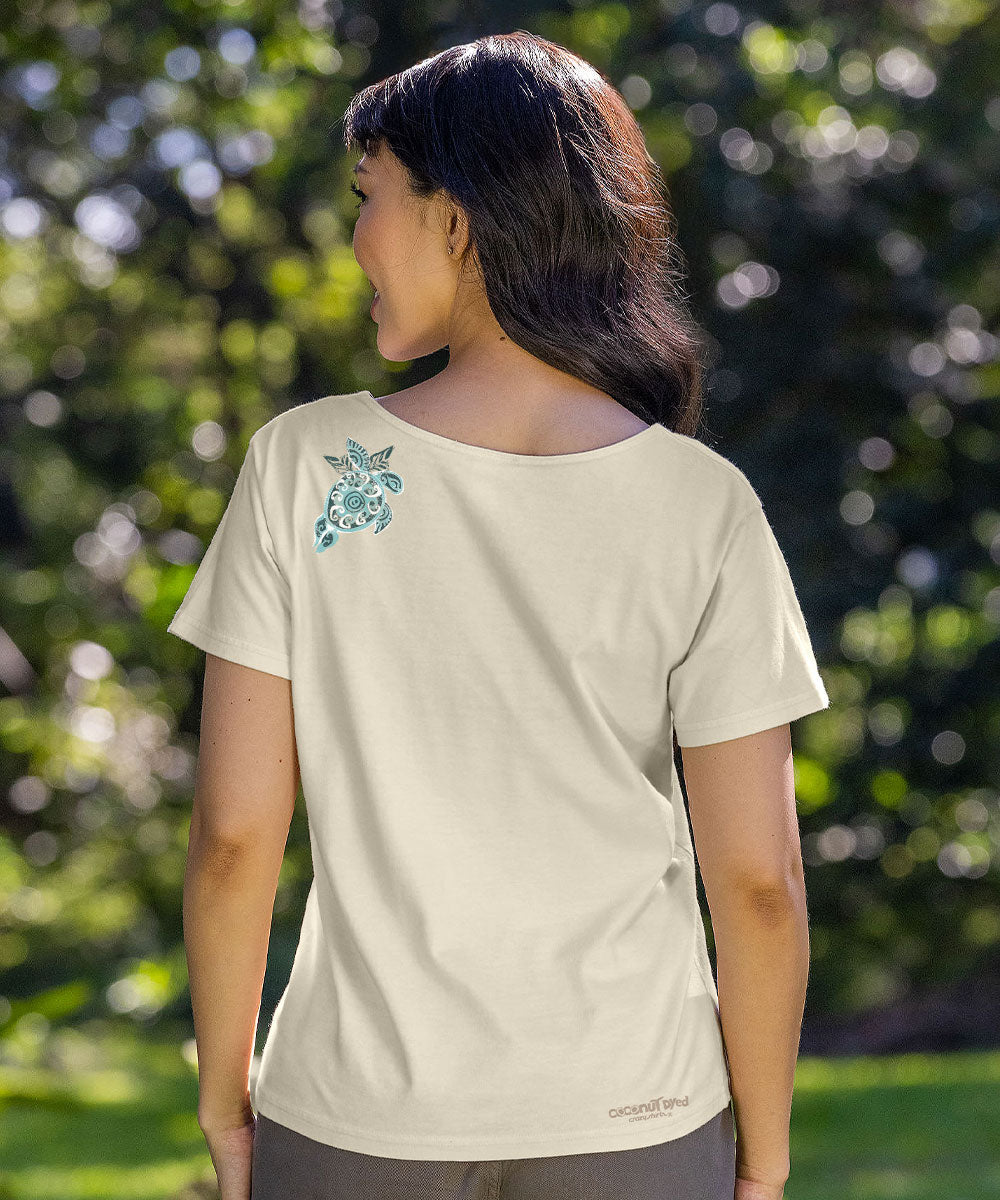 A woman wearing a cream-colored short-sleeve t-shirt with a teal turtle graphic on the left shoulder, photographed outdoors against a blurred green background. View 3