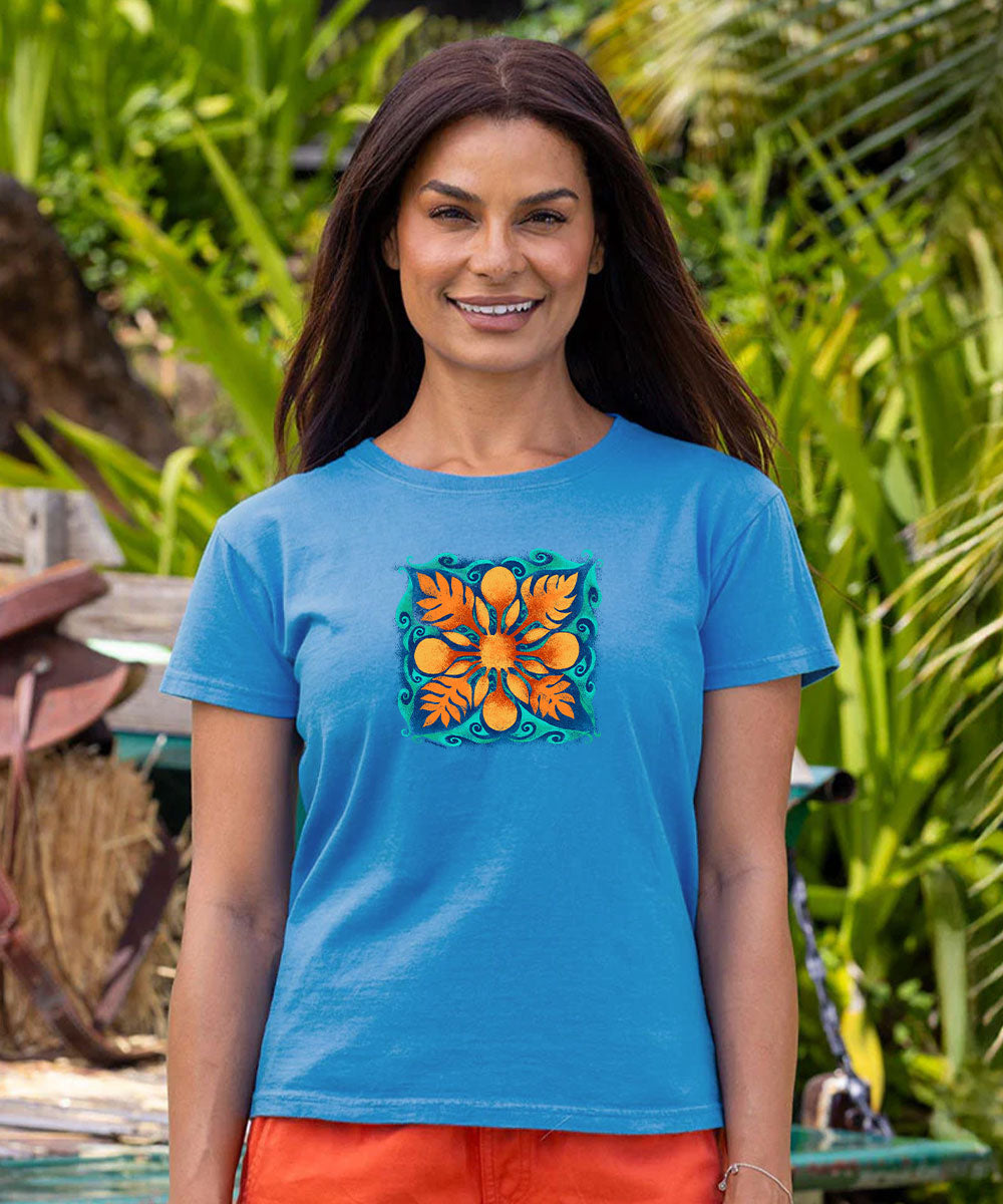 A woman wearing a bright blue short-sleeve t-shirt with a colorful orange and green floral mandala graphic on the chest, set against a blurred background of green tropical foliage. View 2