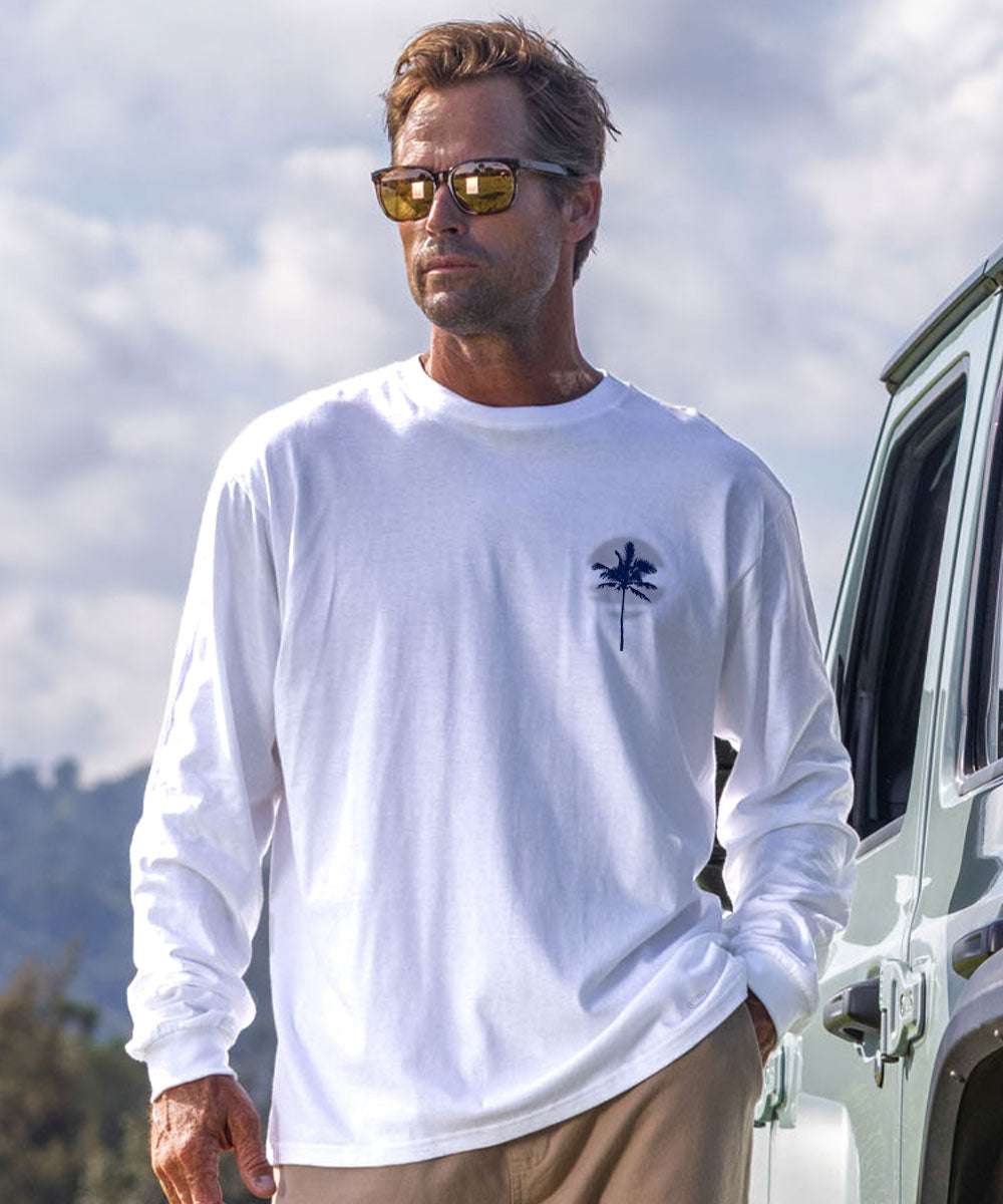 A man wearing a white long-sleeve crewneck t-shirt with a small navy blue palm tree graphic on the left chest, standing outdoors next to a vehicle. View 3