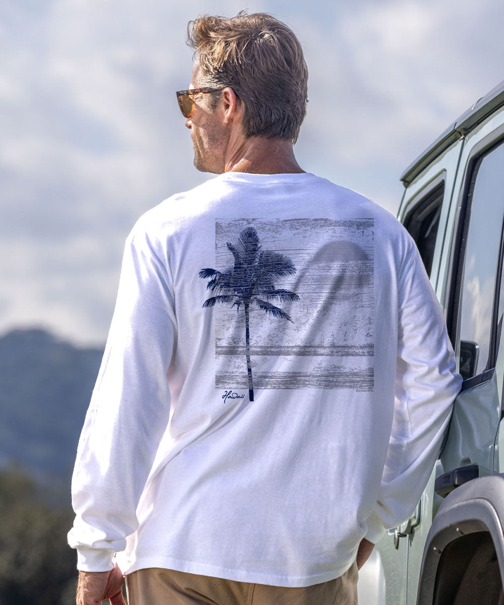 A man wearing a white long-sleeve t-shirt with a dark palm tree graphic on the back, standing outdoors next to a vehicle. View 2