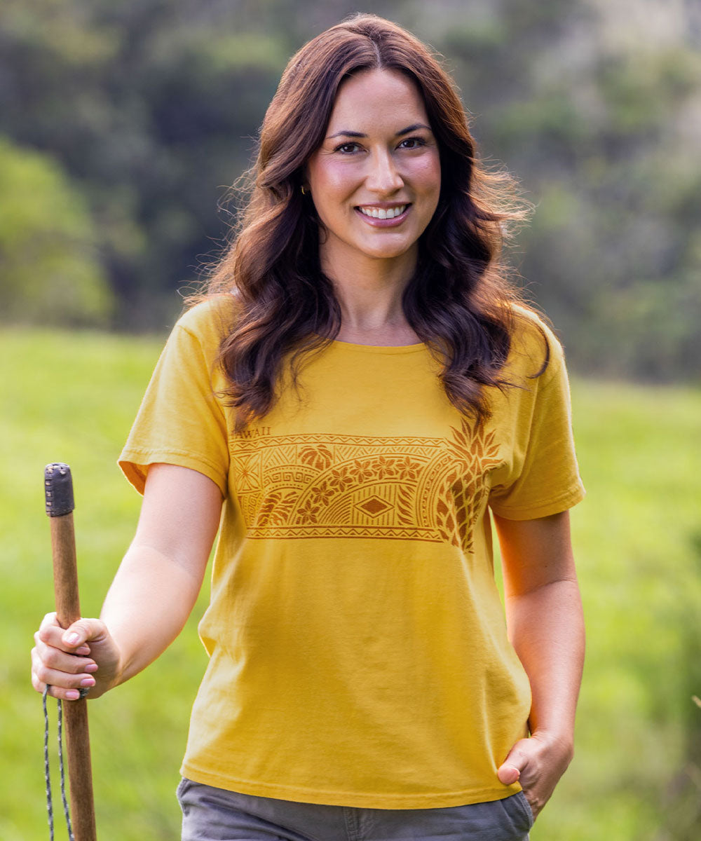 A woman with long brown hair stands outdoors in a grassy field holding a wooden pole, wearing a mustard yellow short-sleeve t-shirt with a brown geometric tribal-style pattern across the chest. View 2