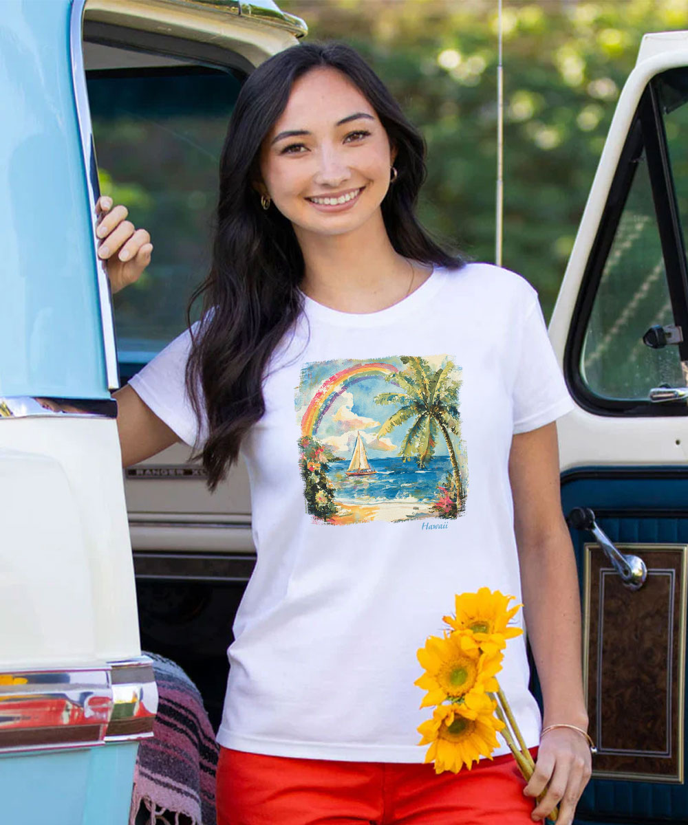 A woman stands outdoors in front of a vintage light blue and white van, wearing a white graphic T-shirt with a tropical beach scene print and bright red shorts, holding yellow flowers. View 2