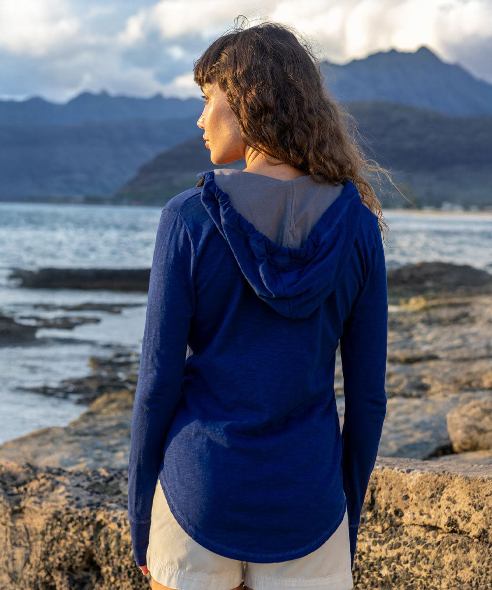 A woman standing on a rocky shoreline with water and mountains in the background, wearing a royal blue hooded long-sleeve top with a gray hood lining and off-white shorts. View 3