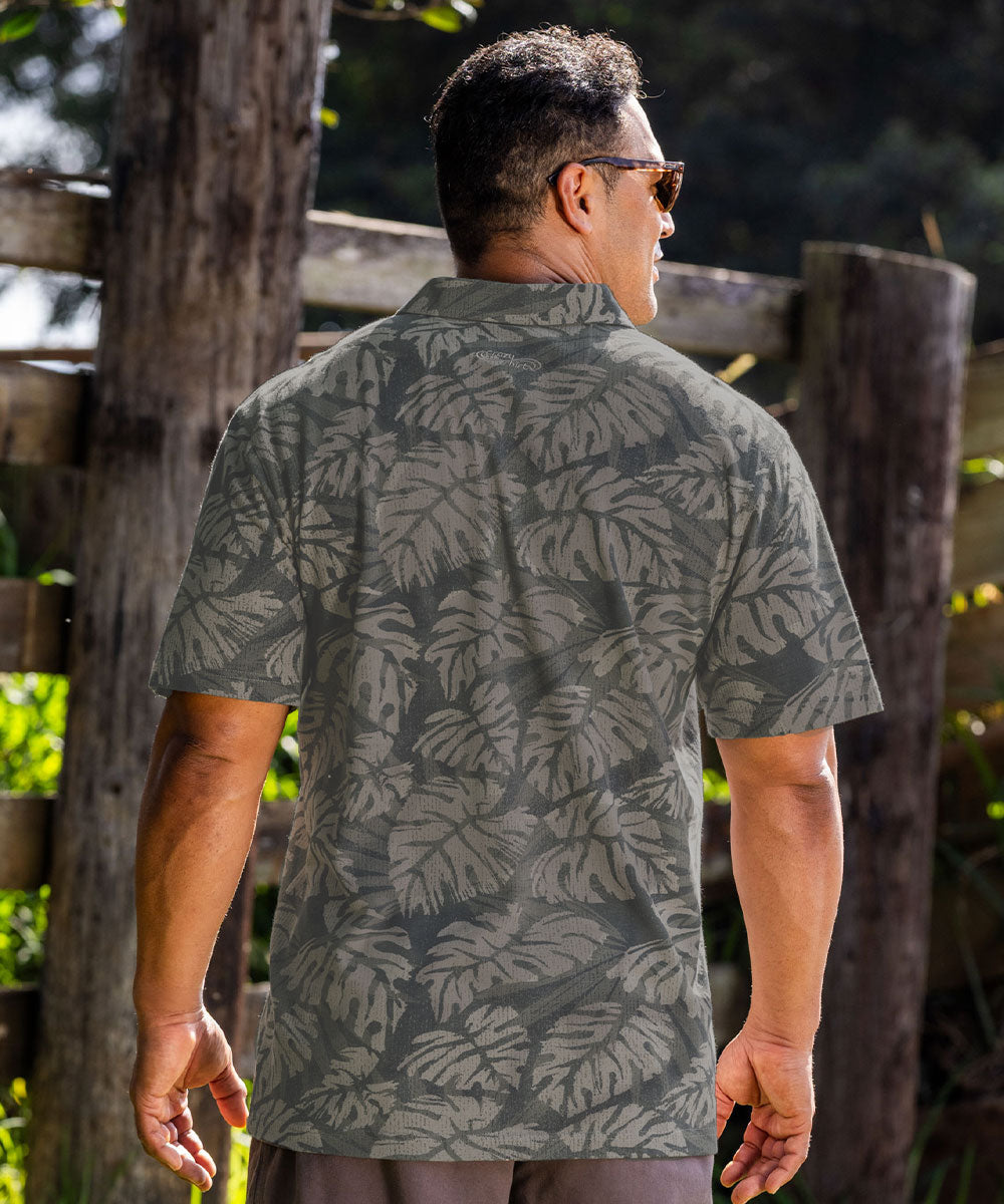A man viewed from behind wearing a short-sleeve button-up shirt with an all-over olive green and gray tropical leaf pattern, set against a rustic wooden fence and green foliage background. View 3