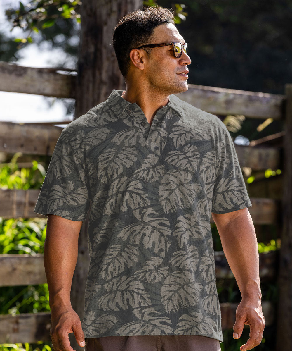 A man wearing a short-sleeve button-up shirt with a gray and olive green tropical leaf pattern, standing outdoors in front of a wooden fence. View 2