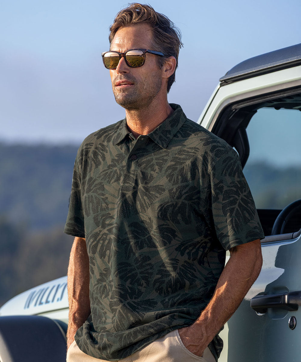A man wearing a dark green short-sleeve polo shirt with a tonal tropical leaf pattern, khaki pants, and sunglasses, standing next to a white vehicle outdoors. View 2