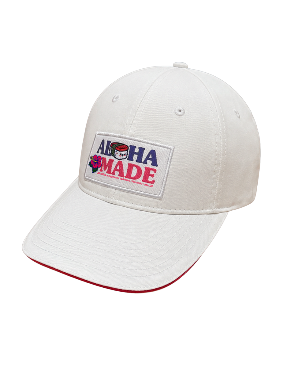 A white baseball cap with a curved brim, featuring a rectangular front patch that reads 'ALOHA MADE' in blue and red lettering with a small graphic. The brim has a thin red piping edge, and the cap is photographed on a clean white background. View 1