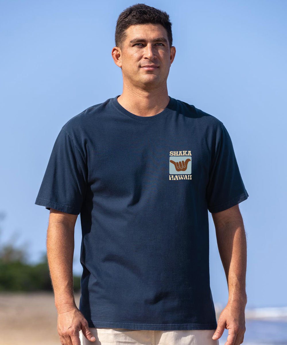 A man wearing a navy blue t-shirt with a 'SHAIKA HAWAII' graphic on the left chest, standing outdoors against a clear blue sky. View 3