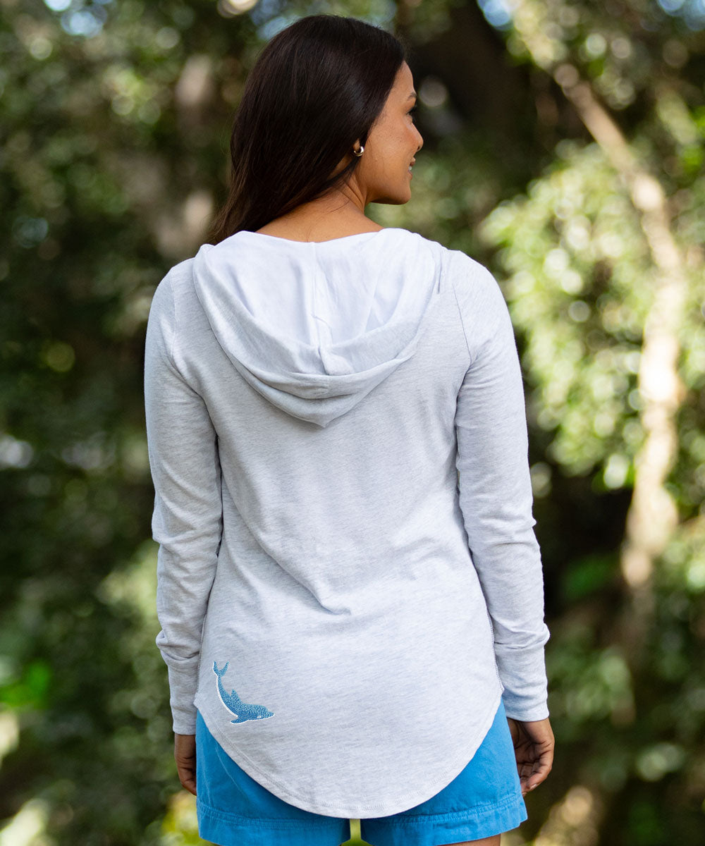 A woman viewed from behind wearing a light gray heather long-sleeve hooded top with a small blue whale graphic near the lower back, set against a blurred green forest background. View 3