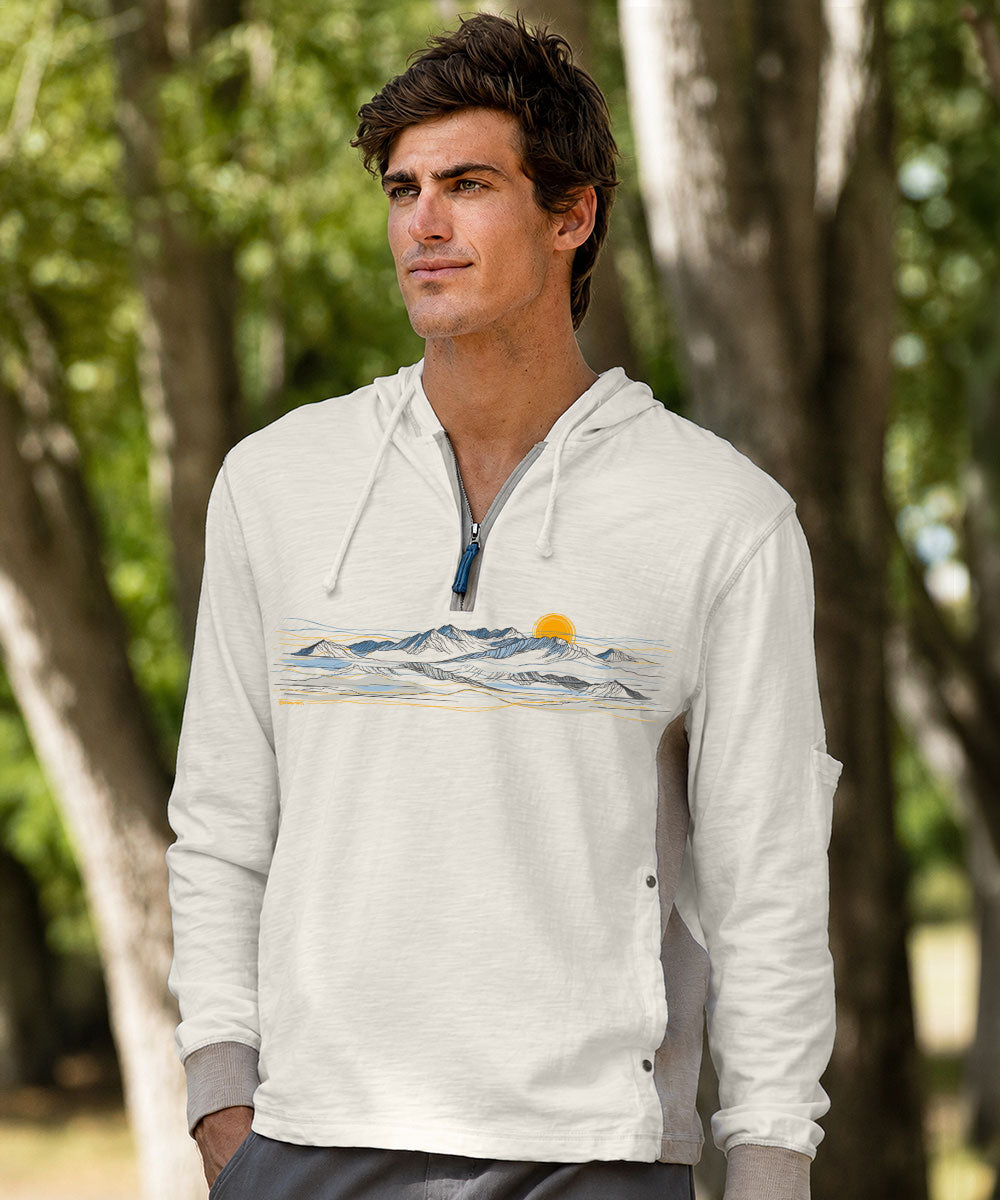 A man wearing a white quarter-zip hoodie with a mountain and sun graphic print on the chest, standing outdoors in front of a blurred background of trees. View 2