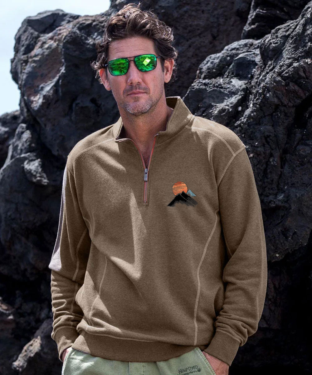 A man wearing a tan quarter-zip fleece pullover with a small mountain and sun graphic on the left chest, standing outdoors in front of dark volcanic rocks. View 2