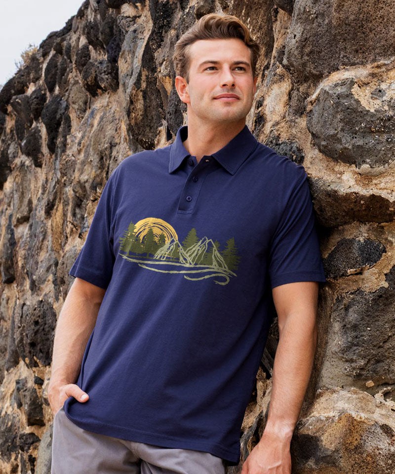 A man wearing a navy blue t-shirt with a gold mountain and forest graphic, sitting outdoors against a stone wall with a blurred green background. View 2