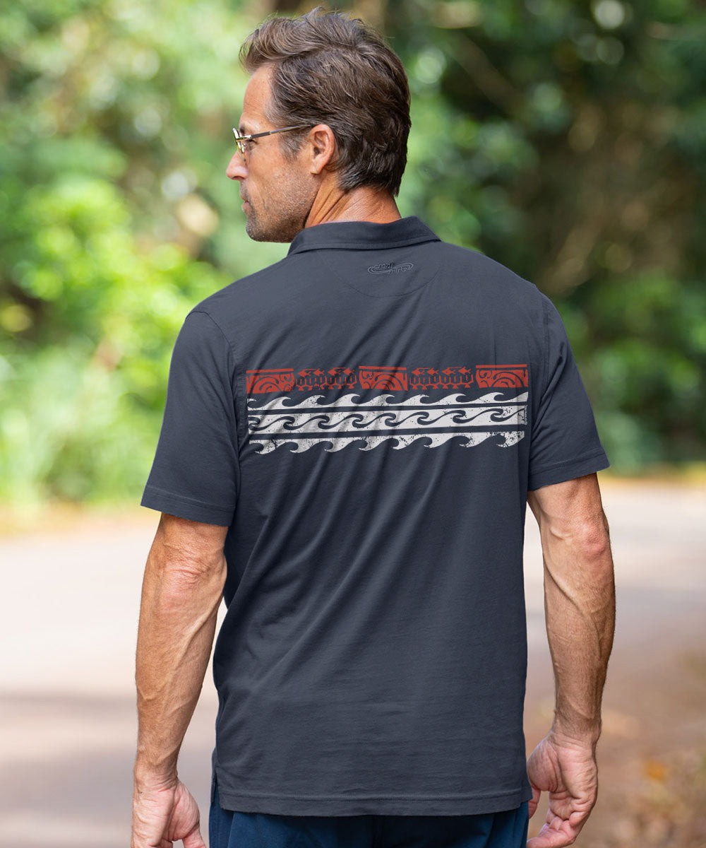 A man viewed from behind wearing a dark charcoal gray polo shirt with a red and white Polynesian-style wave pattern across the upper back, set against a blurred green outdoor background. View 3