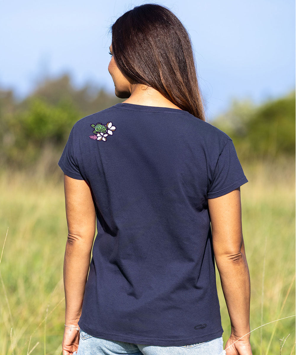 A woman viewed from behind wearing a dark charcoal gray t-shirt with a small green and white turtle graphic on the left shoulder, standing in an outdoor grassy field. View 3