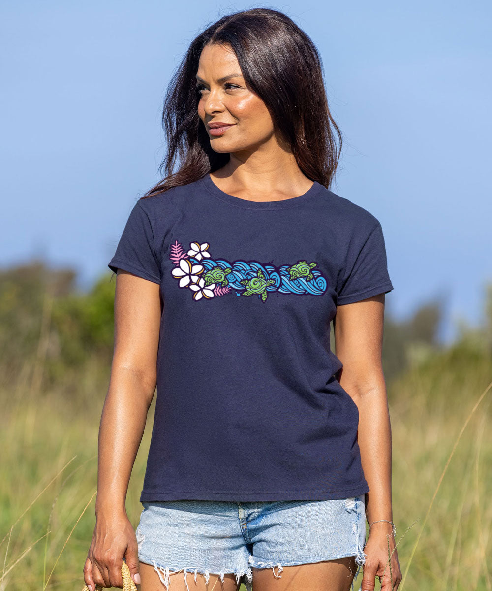 A woman wearing a dark charcoal gray t-shirt with a colorful graphic of green sea turtles and white flowers spelling 'Hawaii', paired with light blue denim shorts, standing in an outdoor grassy field under a blue sky. View 2