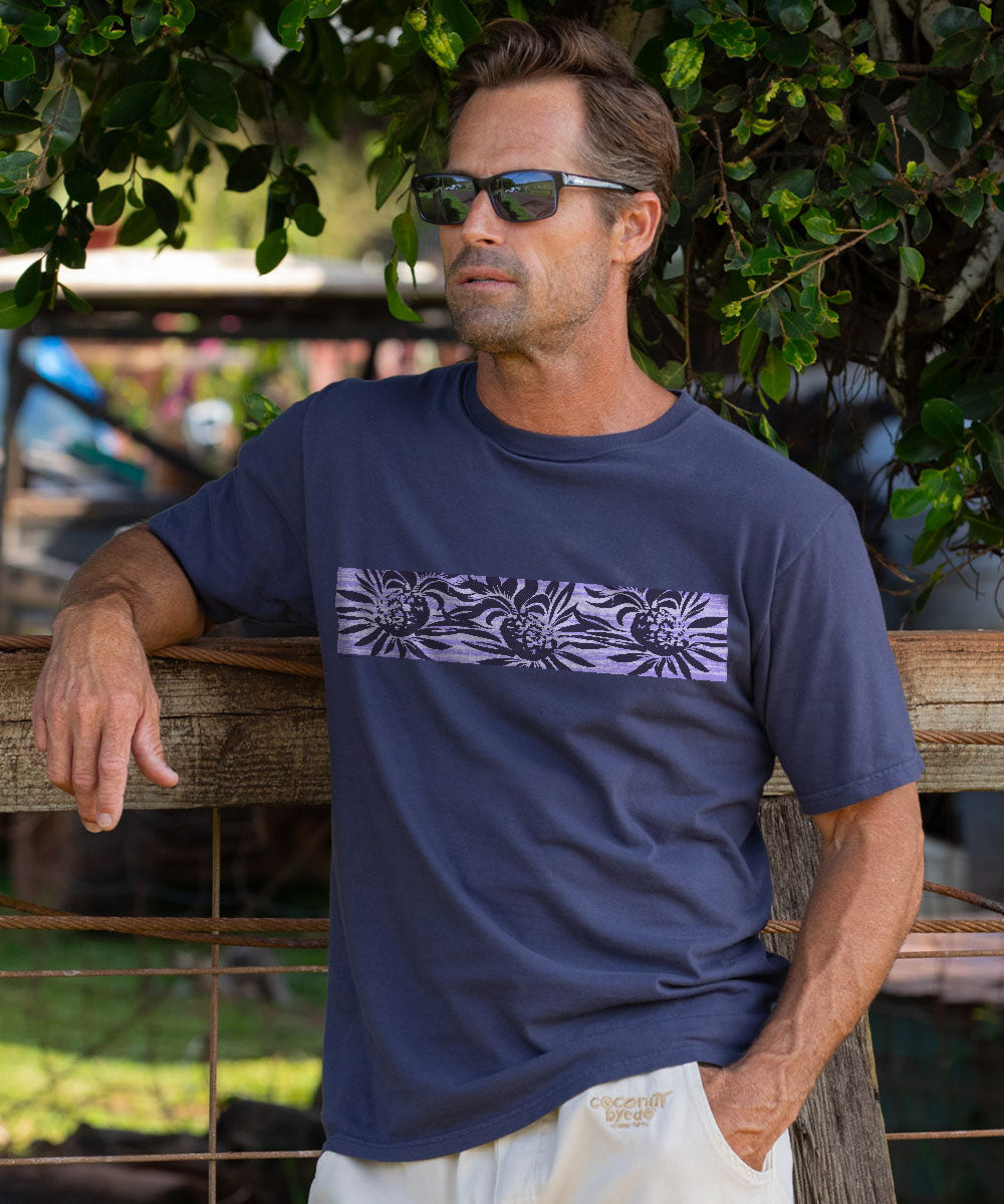 A man wearing a dark charcoal gray t-shirt with a purple and white horizontal floral pattern band, standing outdoors in front of a wooden fence and green foliage. View 2