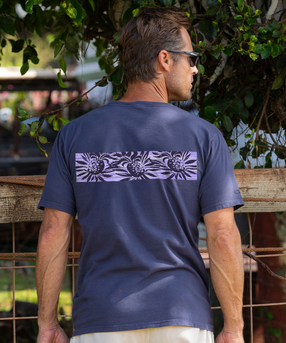 A man viewed from behind wearing a dark gray t-shirt with a horizontal lavender and black floral pattern across the upper back, set against a background of green foliage and a wooden fence. View 3