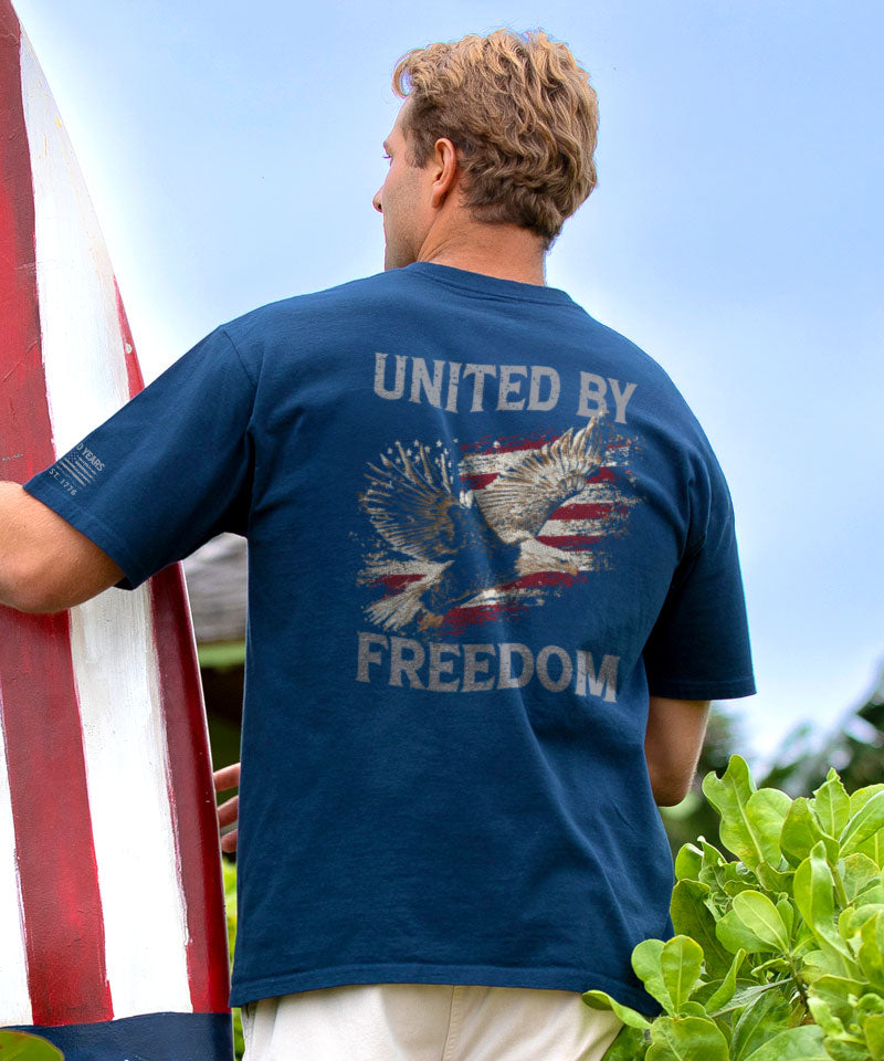 Man wearing a blue t-shirt with 'United by Freedom' design, holding an American flag. View 2