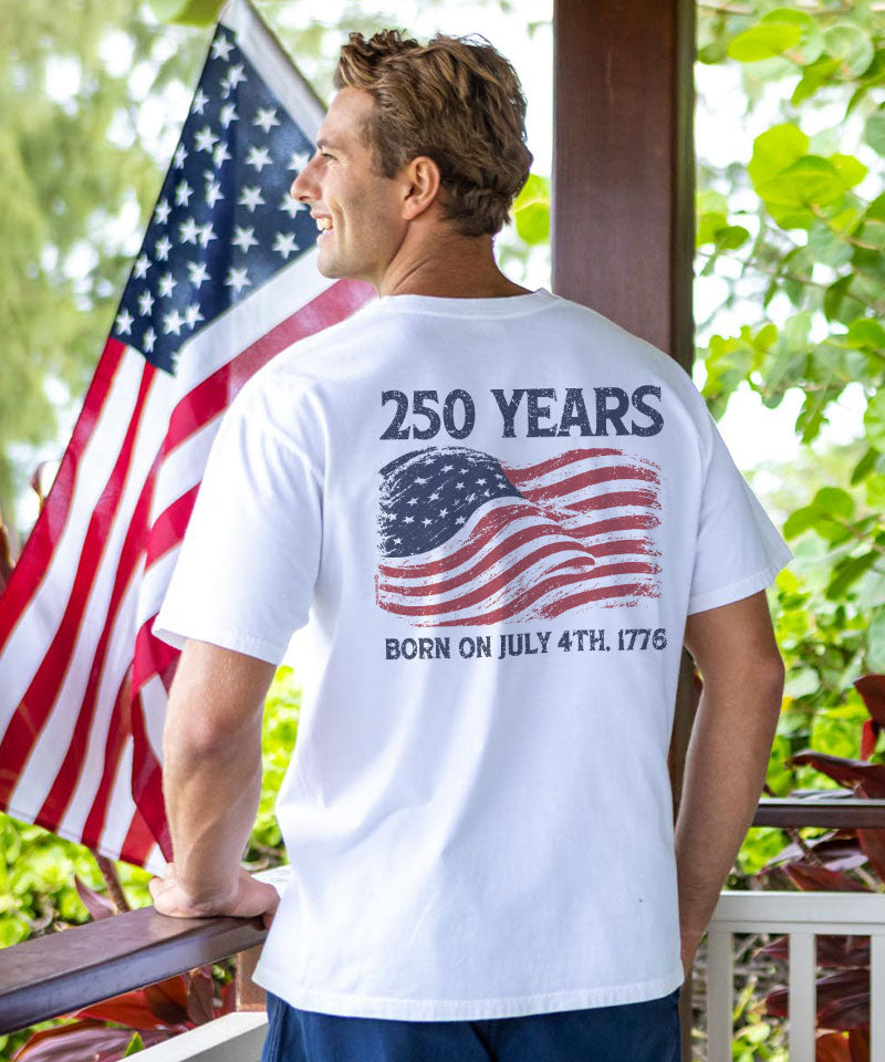 Man wearing a white t-shirt with '250 Years' and American flag design, standing in front of an American flag. View 2