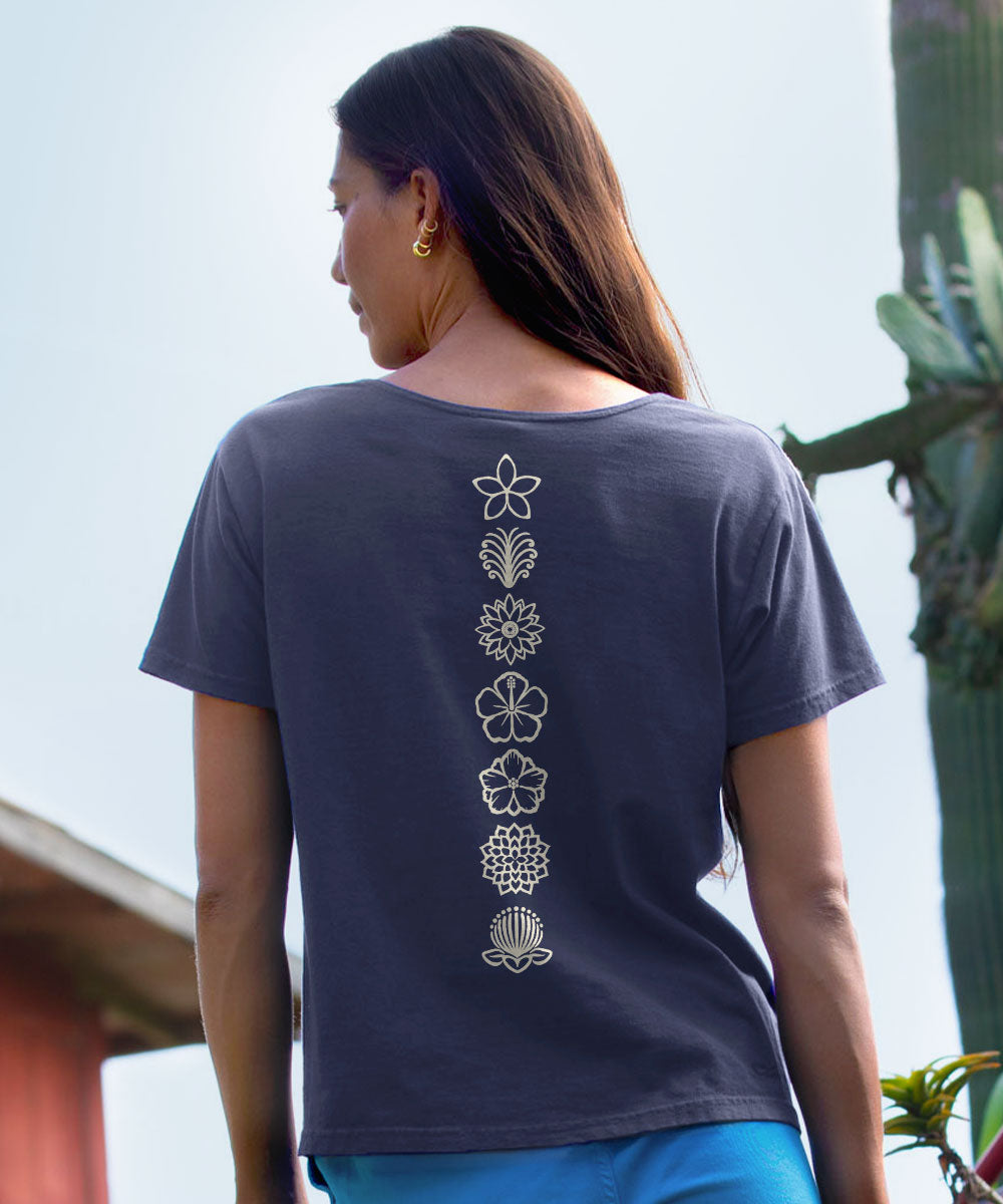 A woman viewed from behind wearing a dark charcoal gray t-shirt with a vertical line of white floral and botanical patterns down the center back, paired with blue pants. View 3