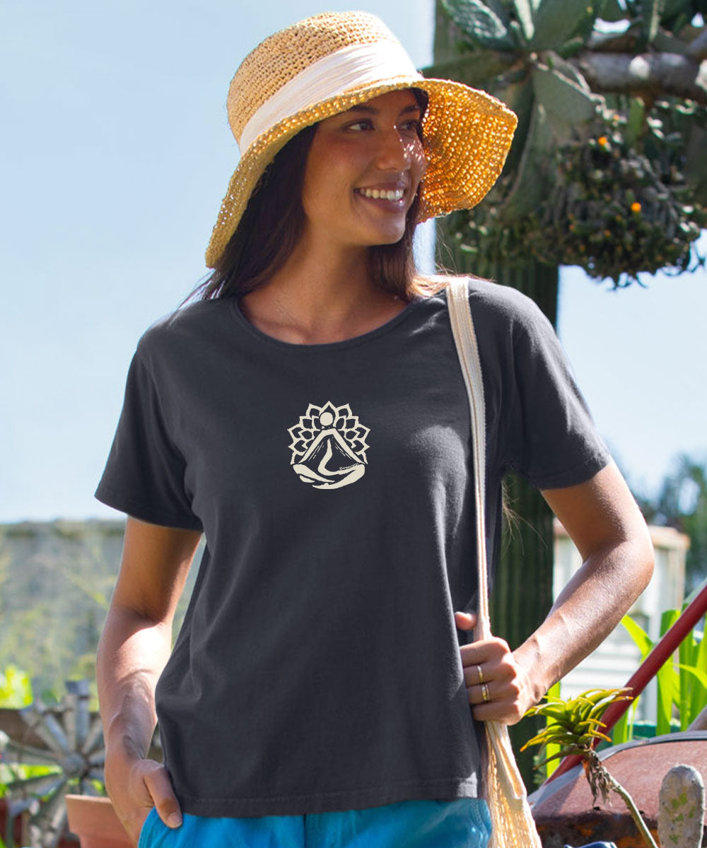 A woman wearing a dark charcoal gray t-shirt with a white sailboat logo on the chest, styled with a straw hat and turquoise pants, photographed outdoors. View 2