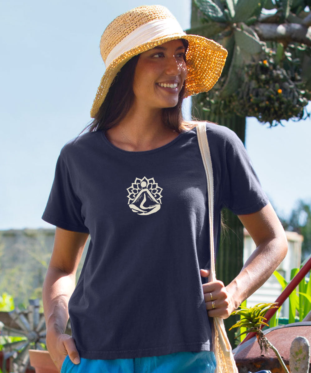 A woman wearing a dark charcoal gray t-shirt with a white sailboat logo on the chest, styled with a straw hat and turquoise pants, photographed outdoors. View 2