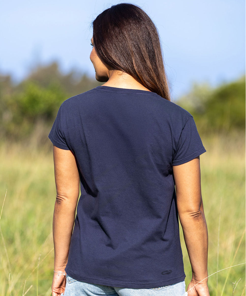 A woman viewed from behind wearing a dark charcoal gray short-sleeve t-shirt and light blue jeans, standing in a grassy field under a blue sky. View 3