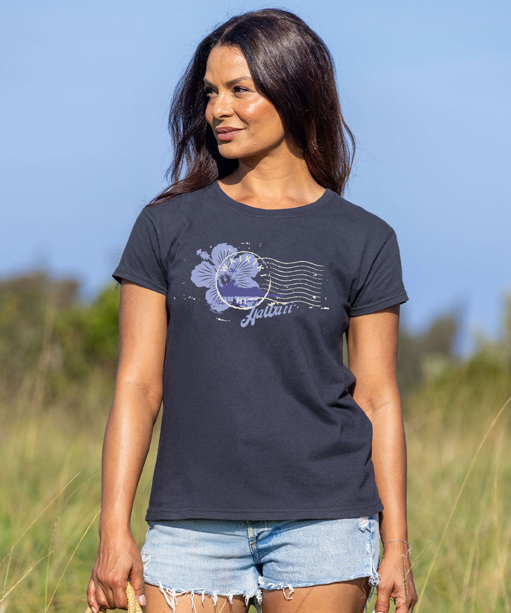 A woman wearing a dark charcoal gray t-shirt with a light purple hibiscus flower and postage stamp graphic, paired with light blue denim shorts, standing in a grassy field under a clear blue sky. View 2