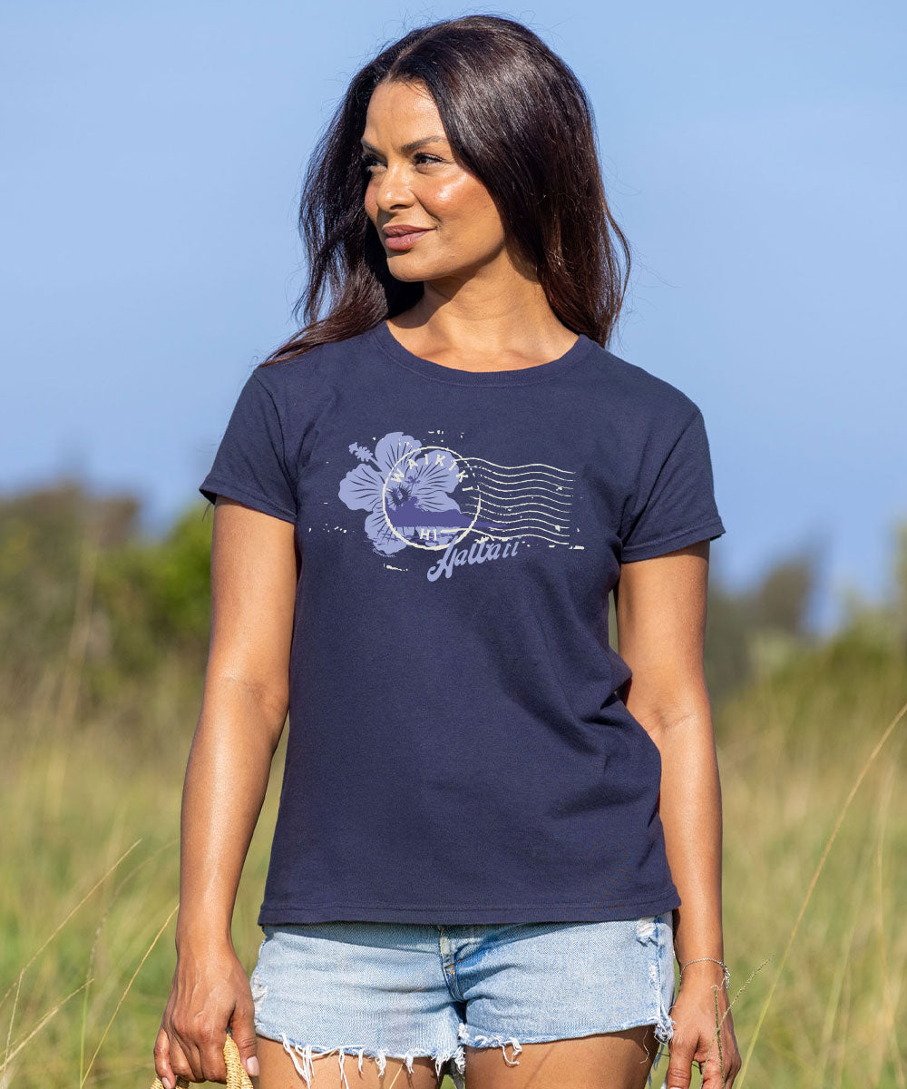 A woman wearing a dark charcoal gray t-shirt with a light purple hibiscus flower and postage stamp graphic, paired with light blue denim shorts, standing in a grassy field under a clear blue sky. View 2