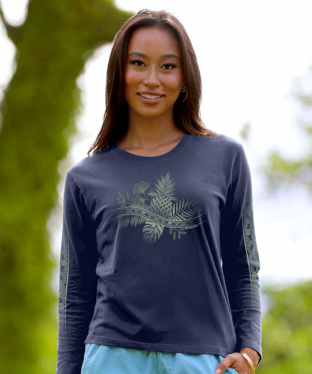 A woman wearing a dark charcoal gray long-sleeve t-shirt with a light green tropical pineapple and palm leaf graphic on the front, set against a blurred green outdoor background. View 2