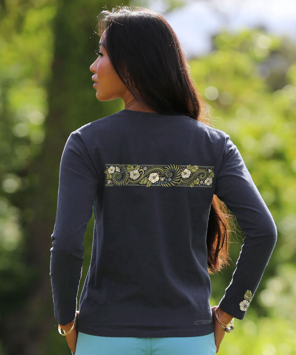 A woman shown from the back wearing a dark navy long-sleeve top with a horizontal floral embroidered band across the upper back and a matching floral detail on the left sleeve, set against a blurred green outdoor background. View 3