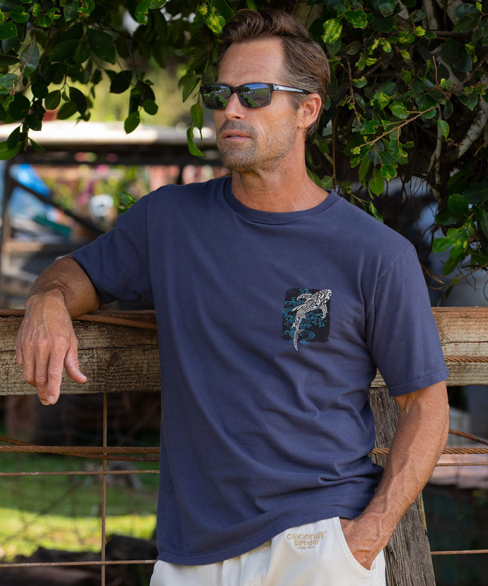 A man wearing a dark gray crewneck t-shirt with a shark graphic on the left chest, leaning against a wooden fence outdoors under green foliage. View 3