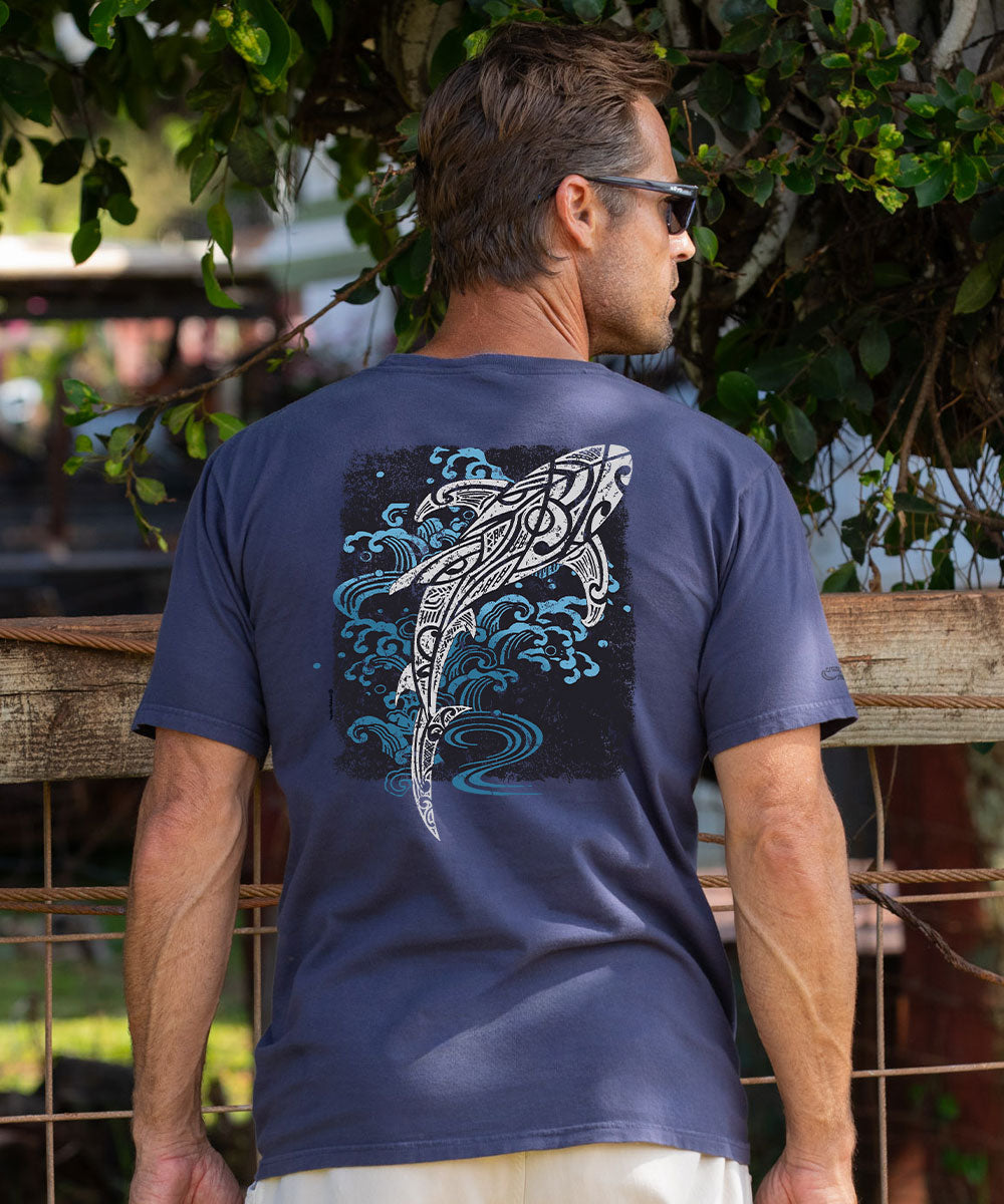 A man wearing a dark gray t-shirt with a large graphic of a stylized shark and blue waves on the back, photographed outdoors in front of a wooden fence and green foliage. View 2