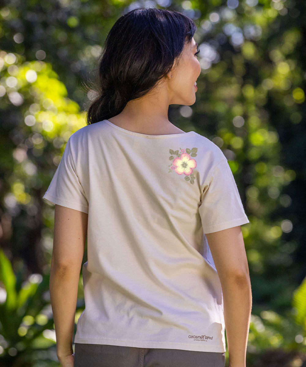 A woman viewed from behind wearing a light beige short-sleeve t-shirt with a small pink and green floral graphic on the upper right shoulder, set against a blurred green outdoor background. View 3