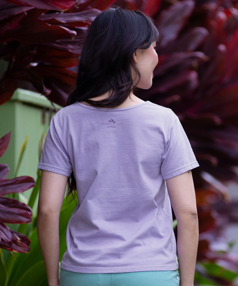 Person wearing a light purple t-shirt with a logo, standing in front of dark red foliage. View 3