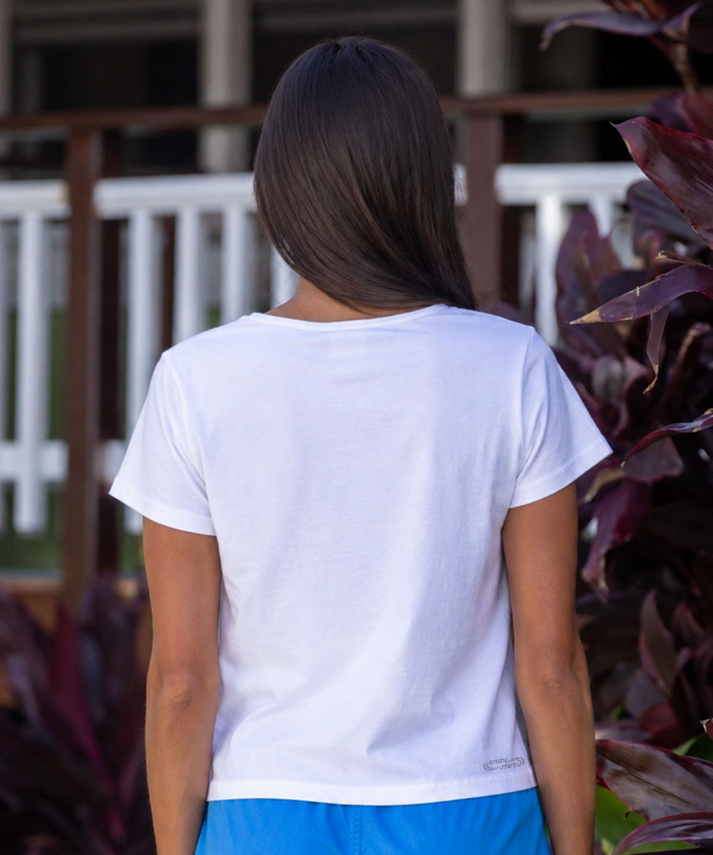 A woman viewed from behind wearing a plain white short-sleeve t-shirt and blue bottoms, standing outdoors in front of a railing and dark purple foliage. View 3
