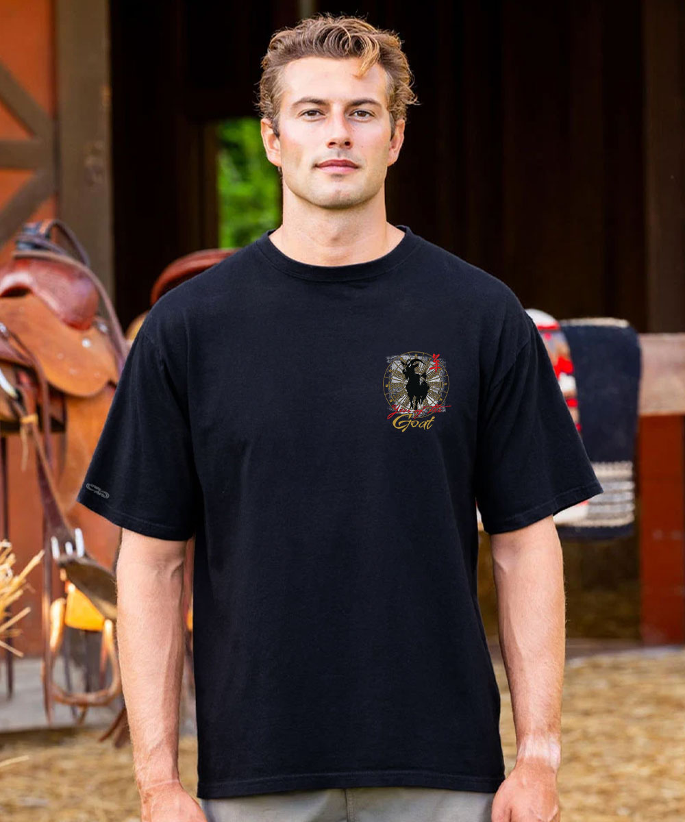 A man wearing a black short-sleeve t-shirt with a small graphic on the left chest, standing in front of a rustic barn interior with saddles and hay visible in the background. View 3