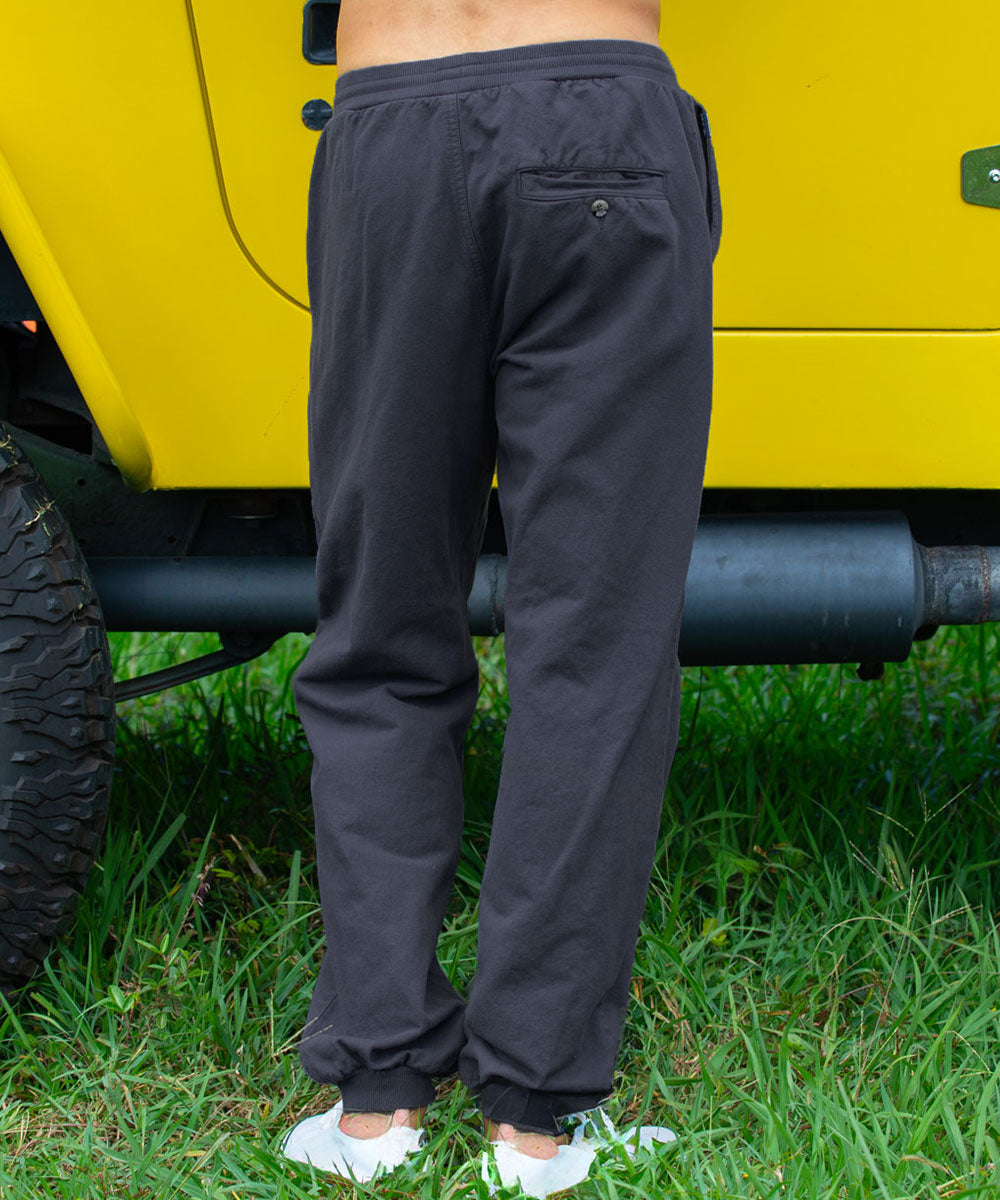 A person standing outdoors on grass beside a bright yellow vehicle, wearing dark charcoal gray jogger-style pants with a back pocket and white sneakers. View 3