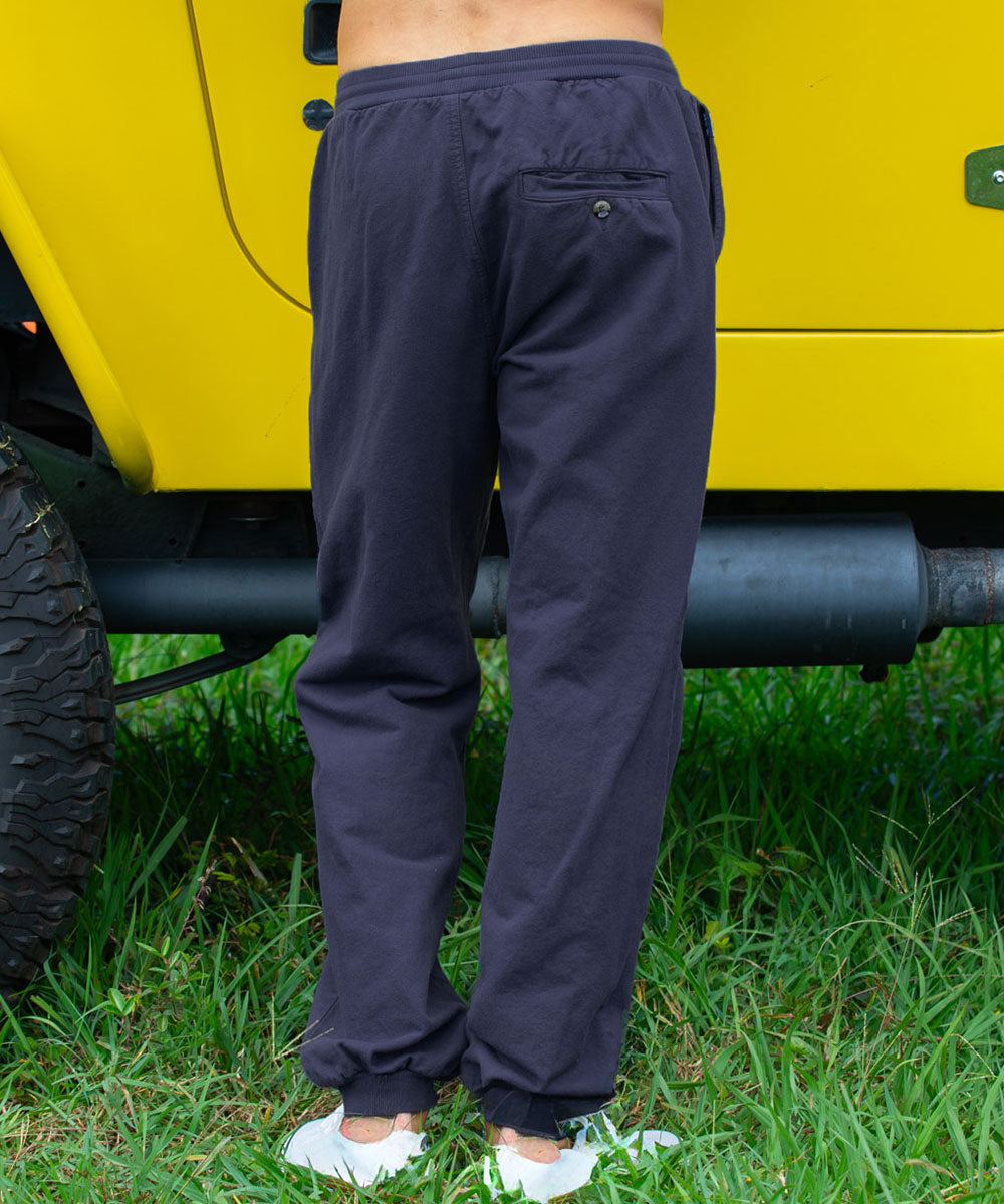 A person standing outdoors on grass beside a bright yellow vehicle, wearing dark charcoal gray jogger-style pants with a back pocket and white sneakers. View 3