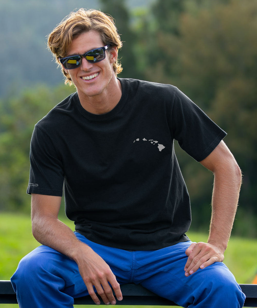 A smiling man wearing a black short-sleeve t-shirt with a small white Hawaii state graphic on the left chest, paired with bright blue pants, sitting outdoors in front of a blurred green background. View 3