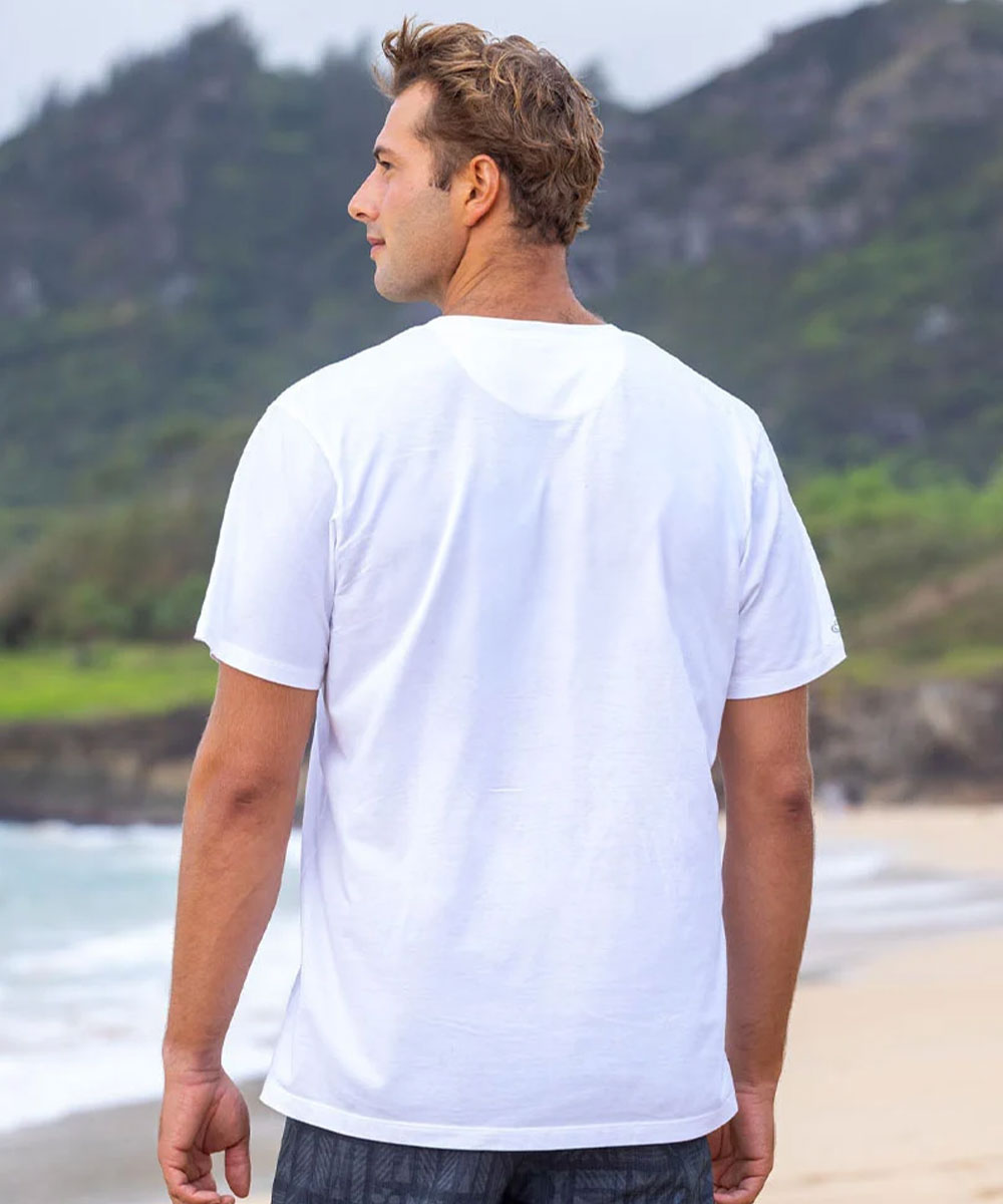 A man standing on a beach, viewed from behind, wearing a plain white short-sleeve t-shirt with ocean waves and green hills in the background. View 3