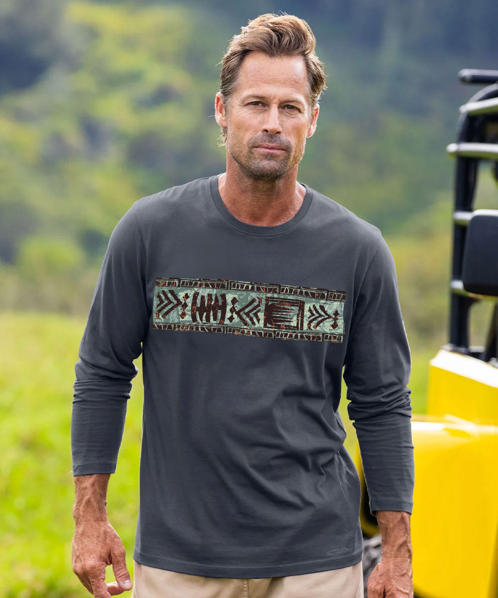 A man wearing a dark gray long-sleeve t-shirt with a horizontal geometric patterned chest graphic, standing outdoors in front of a blurred green background with a yellow vehicle partially visible on the right. View 2
