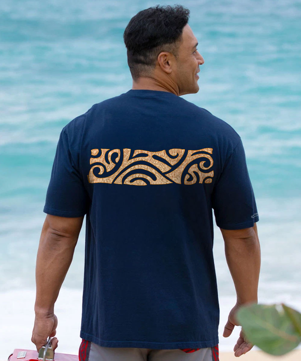 A man viewed from behind wearing a navy blue t-shirt with a tan and black Polynesian-style tribal wave pattern across the upper back, standing on a beach with the ocean in the background. View 3