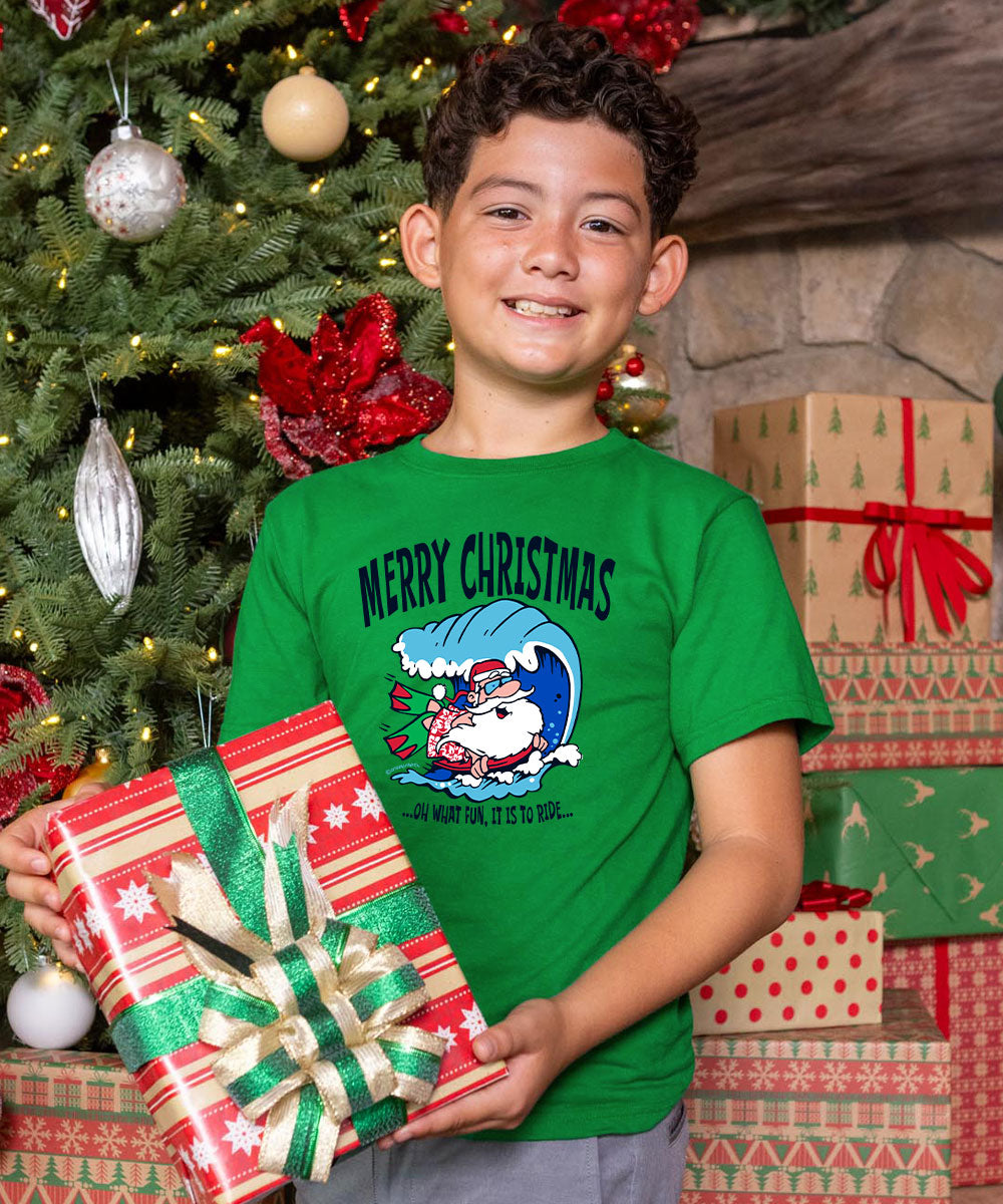 A smiling boy wearing a green Christmas graphic t-shirt stands in front of a decorated Christmas tree holding a wrapped gift. The t-shirt features the text 'MERRY CHRISTMAS' and a cartoon Santa in a blue wave.