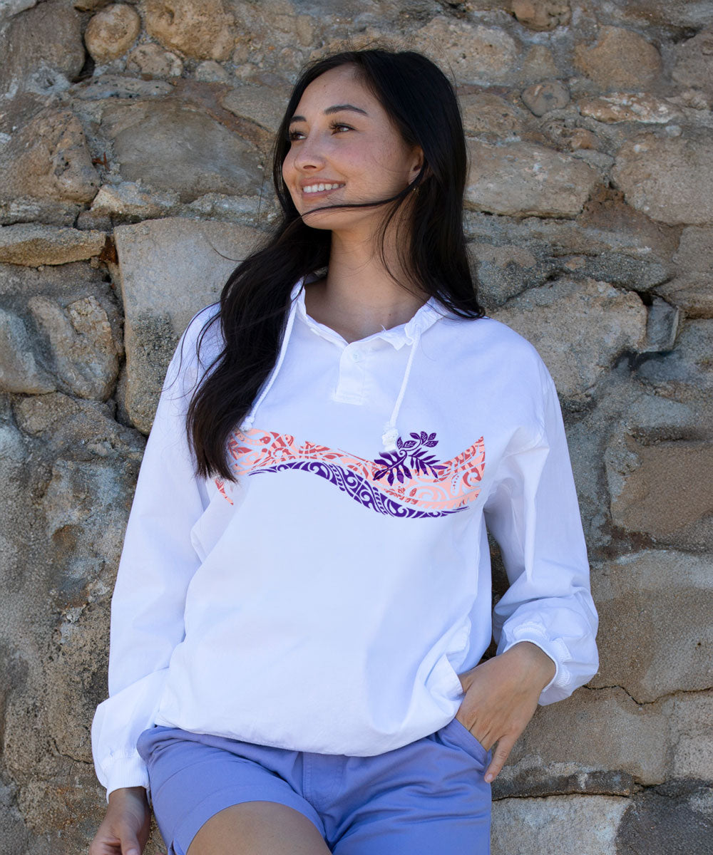 A woman wearing a white long-sleeve hoodie with a pink and purple tropical wave graphic across the chest, paired with light blue shorts, posing against a rustic stone wall. View 2