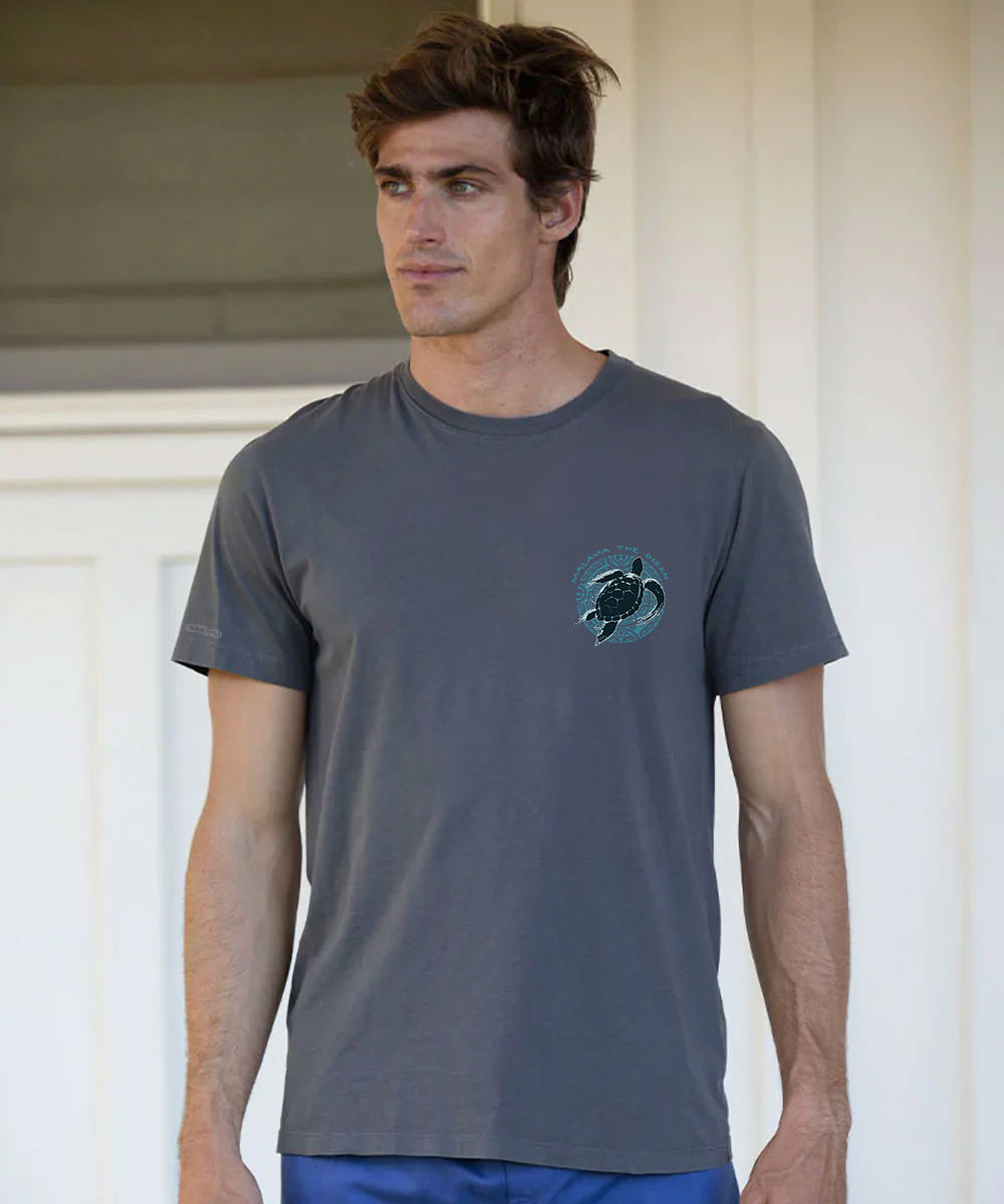 A man wearing a dark gray crewneck t-shirt with a small blue and black sea turtle graphic on the left chest, standing against a white door background. View 3