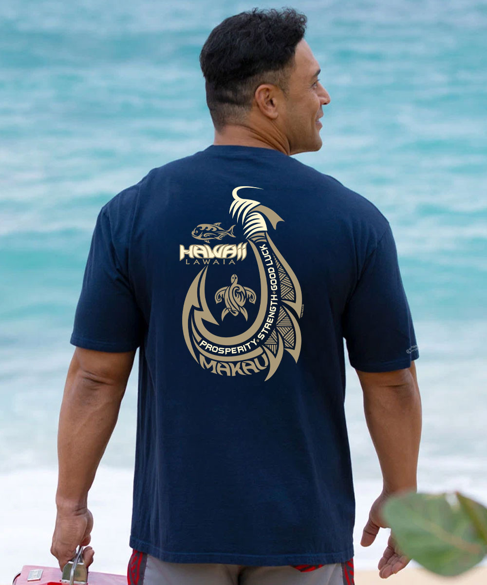 A man viewed from behind wearing a navy blue t-shirt with a large gold Hawaiian graphic on the back, standing on a beach with turquoise ocean water in the background. View 2