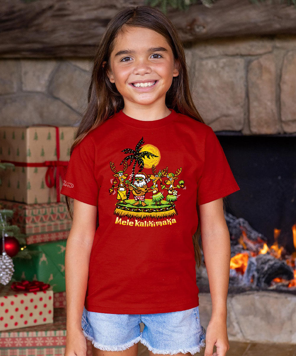 A young girl smiling in front of a fireplace wearing a red graphic t-shirt with a Hawaiian hula dancer design and the text 'Mele Kalikimaka'. View 2